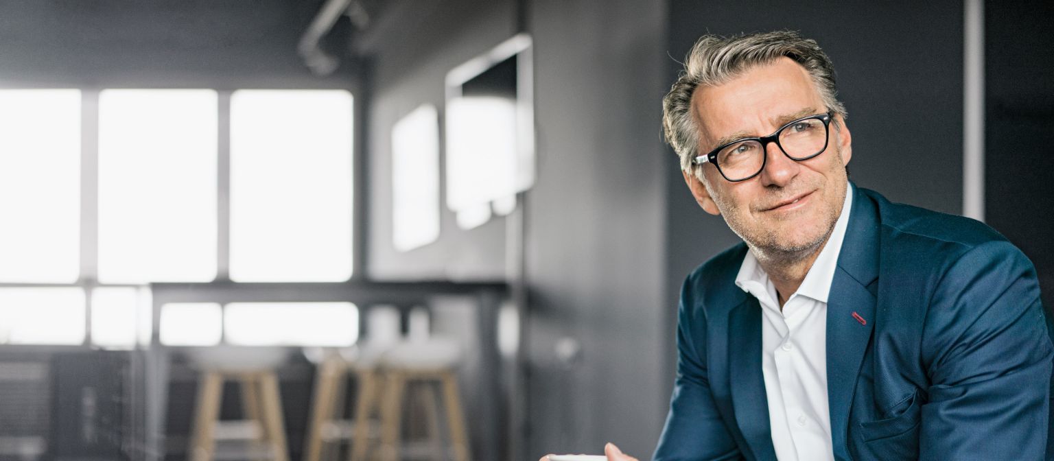 Πορτρέτο ενός καθιστού ώριμου επιχειρηματία που φοράει μπλε κοστούμι και κρατάει μια λευκή κούπα καφέ.